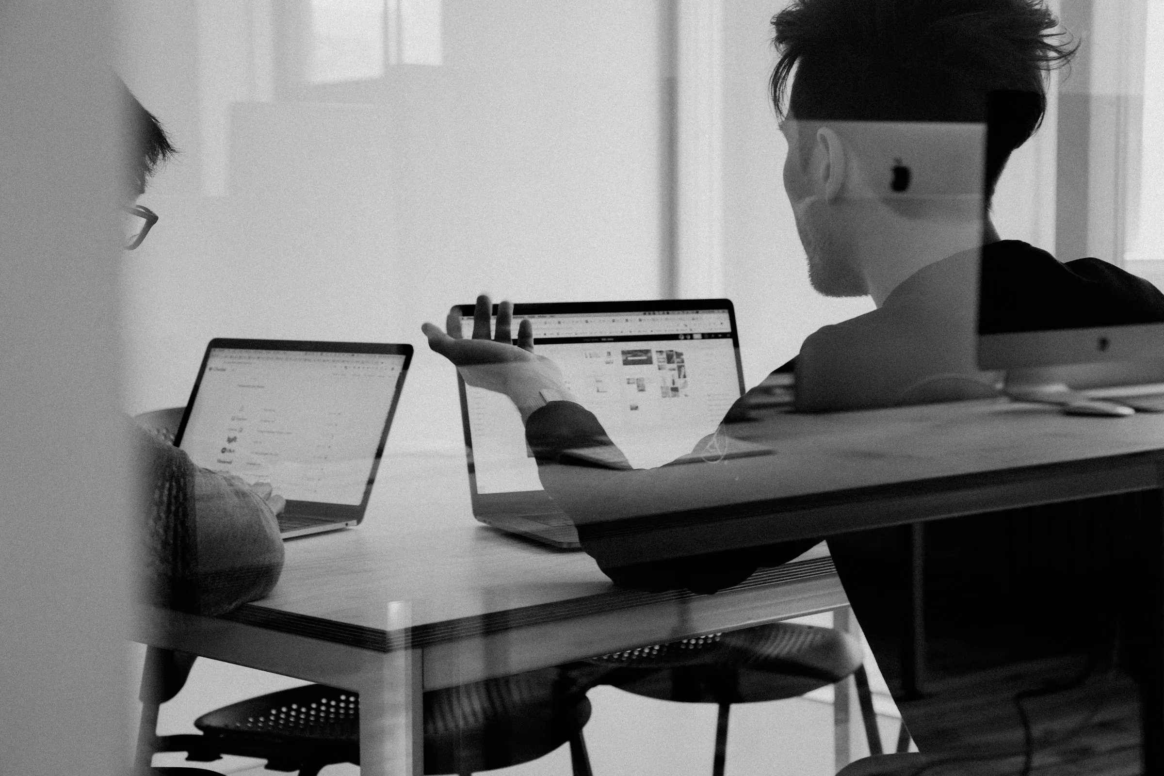 Two colleagues discussing work at laptops, seen through glass (Unsplash free license)