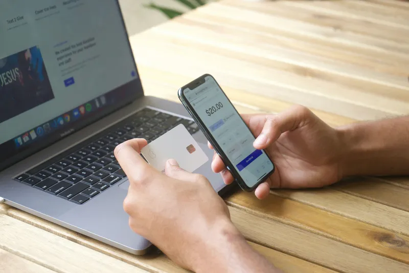 Person using a banking app on a smartphone with a payment card next to a laptop